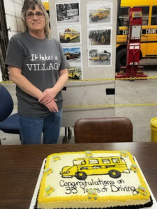 A woman stands behind a cake that's designed with icing in the shape of a school bus and the words Congratulations on 35 Years of Driving