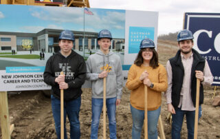 A picture of Johnson Central High School students Brandon Holbrook, Hunter Burchett, Constance Martin and Nick Hardin standing in front of a construction sign and holding on to shovels, wearing construction hats.