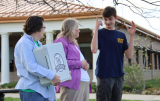 Three people stand as a teen communicates through sign language and a woman holds a box with the words 25 Years written on it