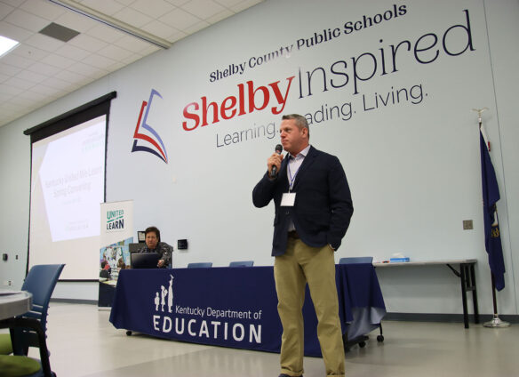 Education Commissioner Jason E Glass stands at the front of a room and speaks into a handheld microphone. A table behind him reads Kentucky Department of Education