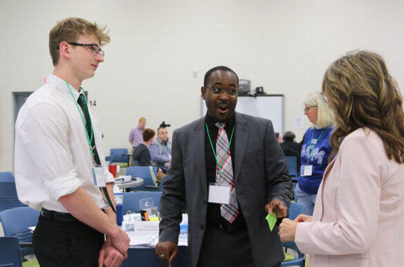 Three people stand talking in a conference room. The person in the middle excitedly looks to the person on their right.