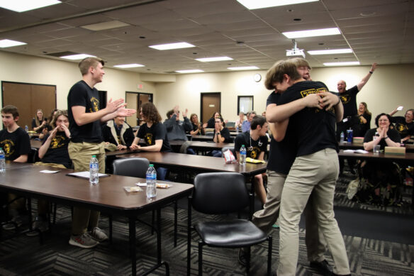 Two students hug as several other people cheer and clap their hands