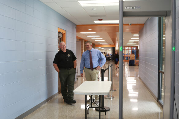 Two men stand by a door and smile as a metal detector stands in front of them