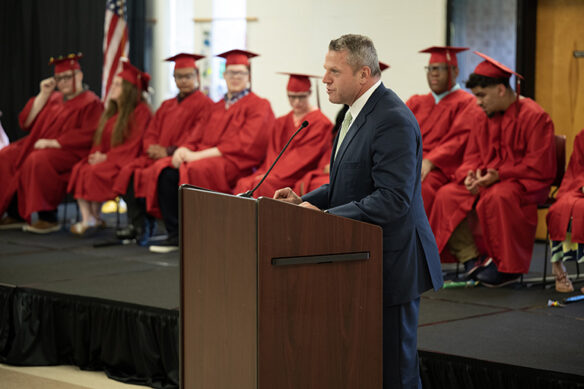 Picture of Jason E. Glass standing at a podium and speaking with the students sitting behind him in their caps and gowns.
