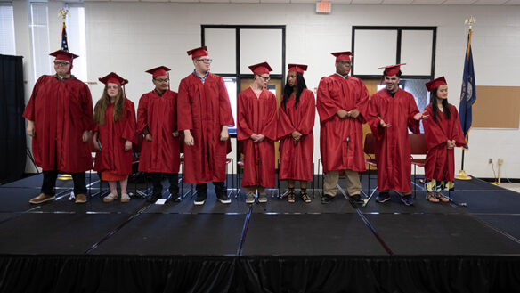 Picture of nine students in graduation caps and gowns standing on a stage during a ceremony.