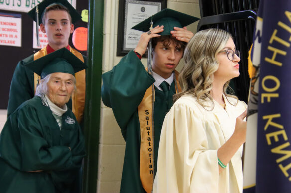 Three people in green graduation cap and gowns stand in line with a girl in front of them who is wearing a white robe and holding the state flag of Kentucky. One of the graduating students adjusts his hat.
