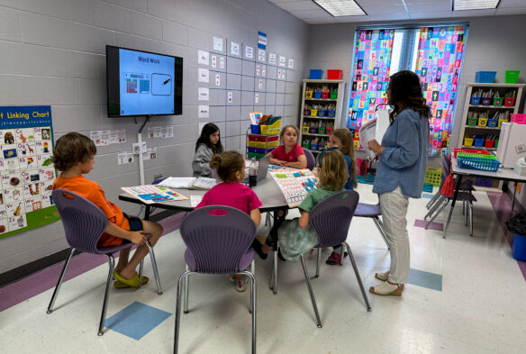 A teacher stands over a desk with six students and instructs them on the screen through "Word Work"