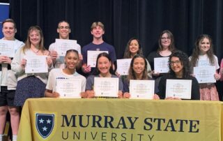 A group of students hold pieces of paper to show they have committed to Murray State University for teaching