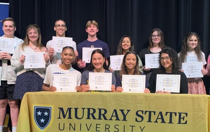 A group of students hold pieces of paper to show they have committed to Murray State University for teaching