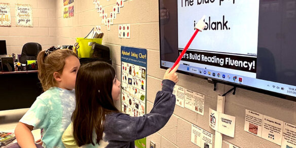 A picture of two girls in a classroom using a long stick to point to words on a screen.