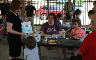 An adult holds a book that she is about to give to a child participating in a summer reading program. The student is also eating lunch.