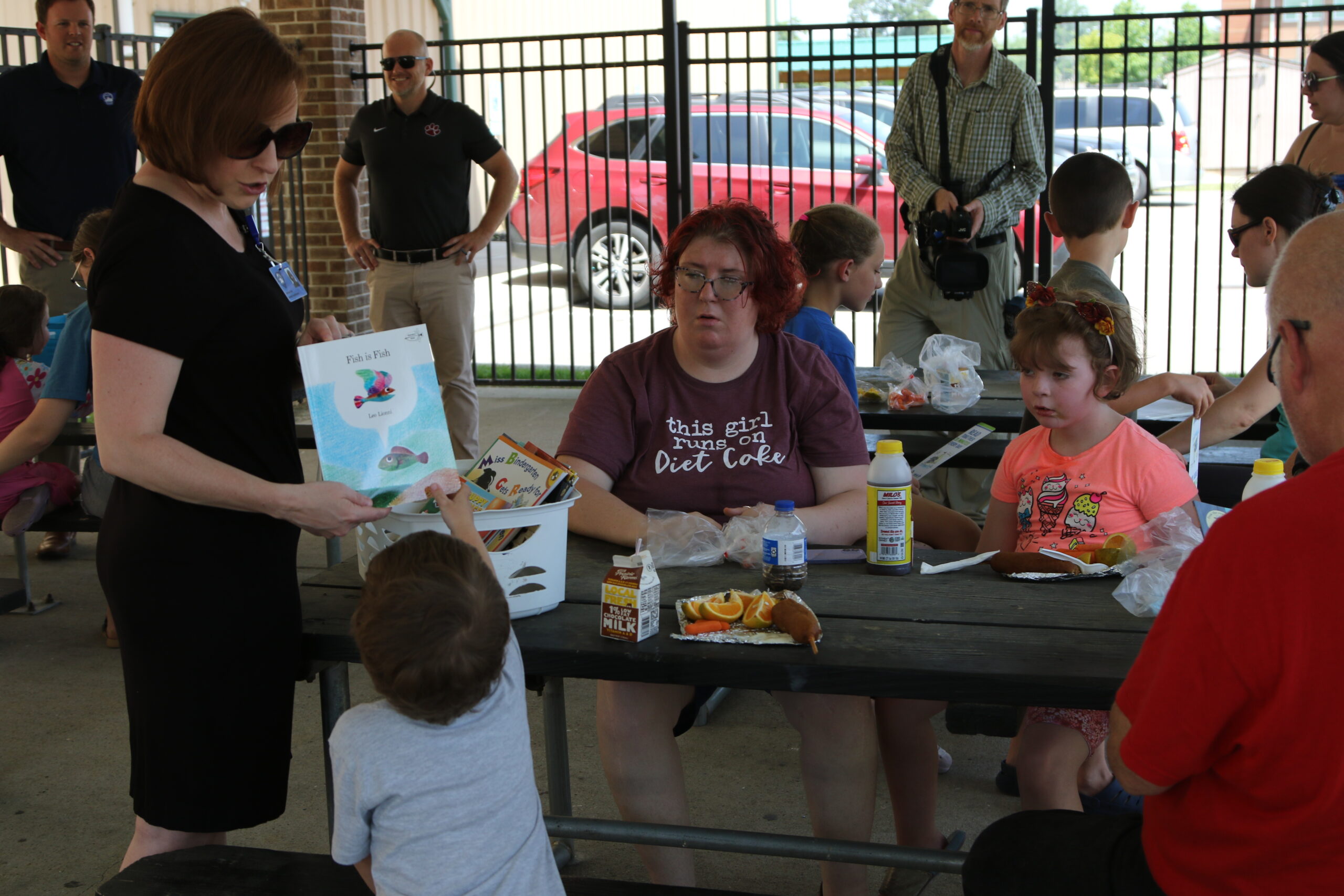 An adult holds a book that she is about to give to a child participating in a summer reading program. The student is also eating lunch.