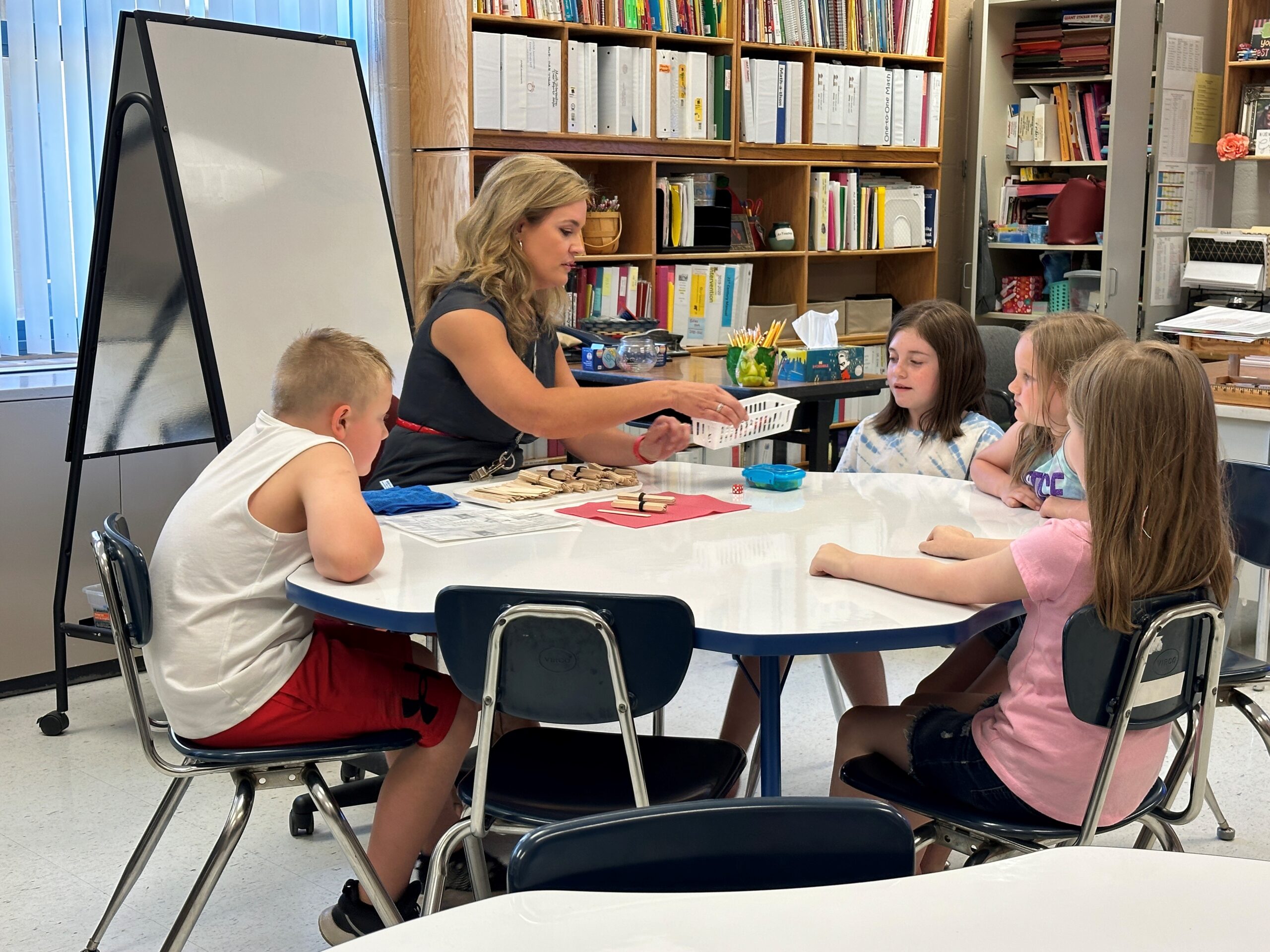 A teacher sits at a table with four students. She is handing out piece to a math game.