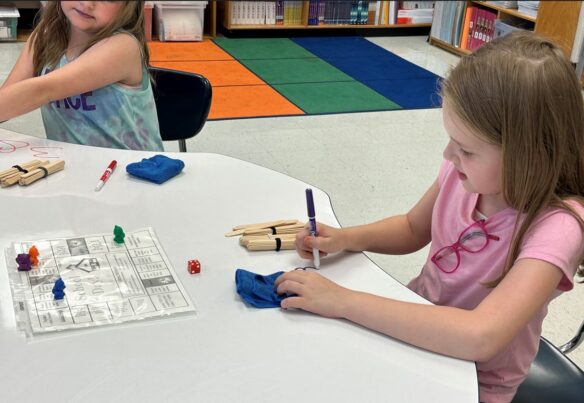 a female student is using a dry erase marker to draw a number on a table as part of a math game inside a classroom.