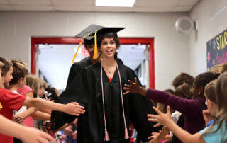 A woman in a graduation gown walks down a hallway, hands extended to greet kids in the hallway