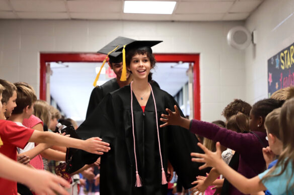 A woman in a graduation gown walks down a hallway, hands extended to greet kids in the hallway