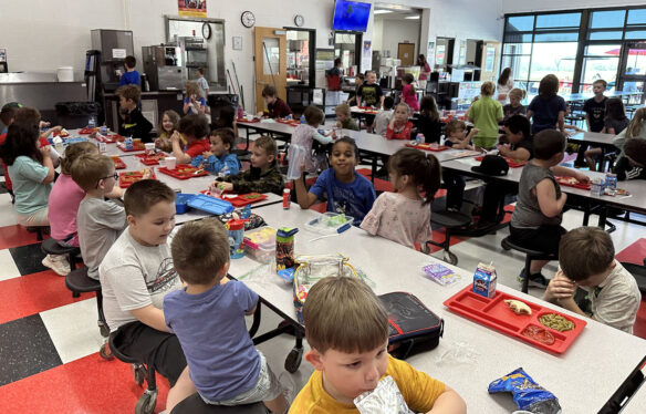 Picture of a school lunchroom filled with young students eating lunch at tables and talking to each other.
