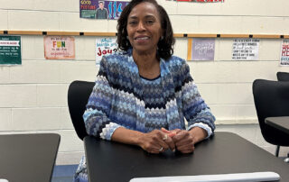 A woman is seated in a classroom at a desk. She is smiling.