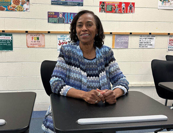 A woman is seated in a classroom at a desk. She is smiling.