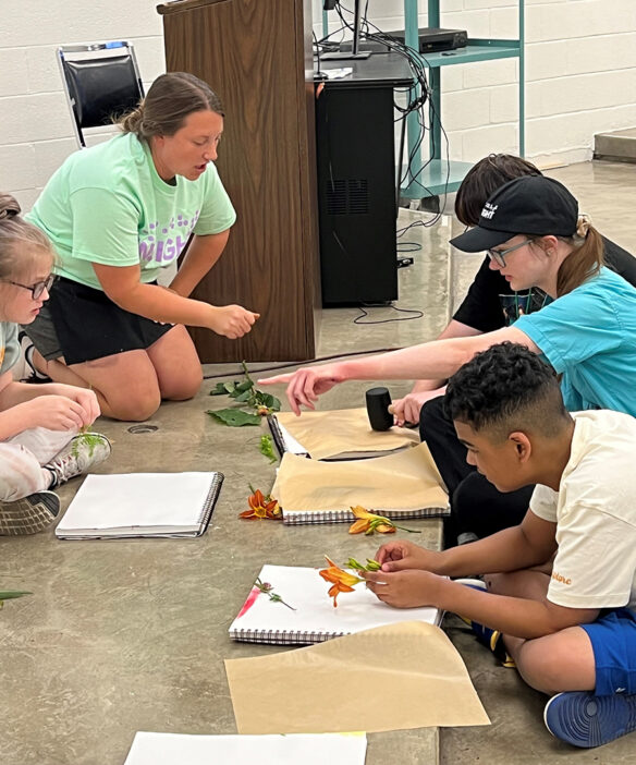 Picture of students sitting on a floor, making an art project with flowers while an instructor talks with them.