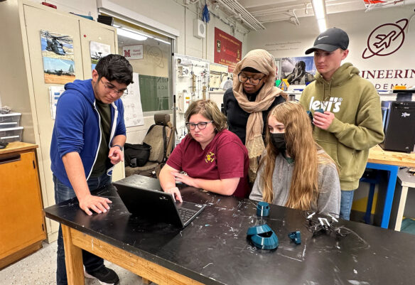 Picture of five high school students sitting and standing around a computer with a drone lying on the table in front of them.