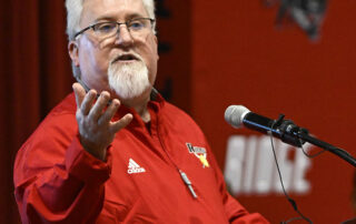 A picture of David McCoy speaking at a podium with his arm stretched out.