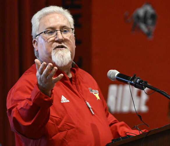 A picture of David McCoy speaking at a podium with his arm stretched out.