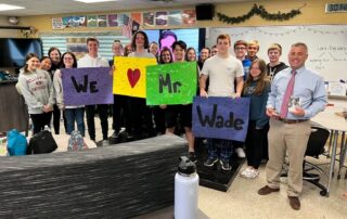 Picture of Mark wade standing in front of a classroom with a bunch of high school students holding signs that say we love Mr. Wade.