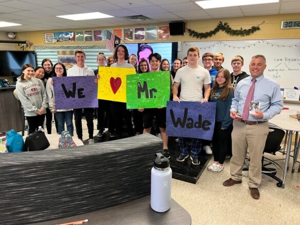 Picture of Mark wade standing in front of a classroom with a bunch of high school students holding signs that say we love Mr. Wade.