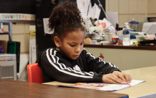 A female elementary student is seated at a desk. She is reading Braille.