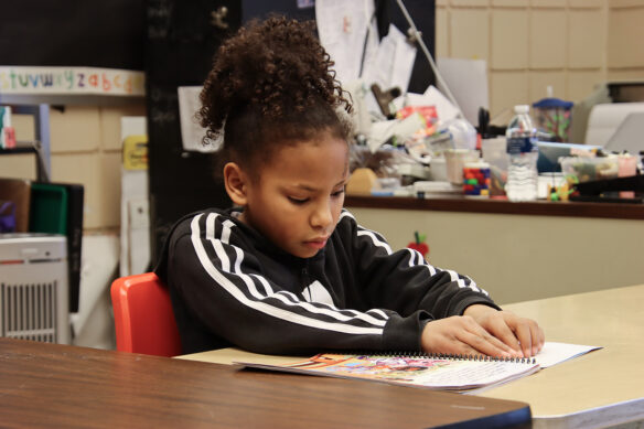 A female elementary student is seated at a desk. She is reading Braille.