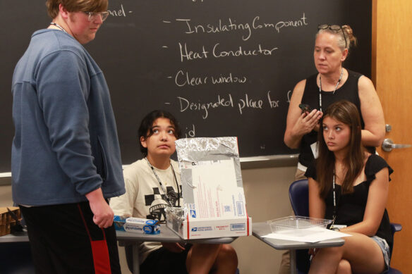A group of three students in a classroom talk to instructor Marie Price about their project, which is a small box coated in aluminum foil.