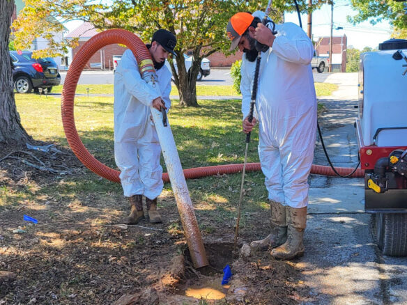 Two men stand while one is holding a hose pointed at a hole in the ground that has dirty water in it. The hose is attached to a tank.