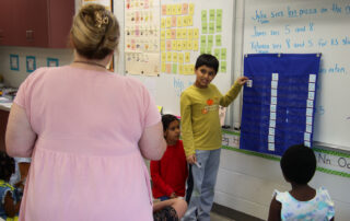 A child holds up a card that has the number 21 on it as he looks at a teacher in a classroom