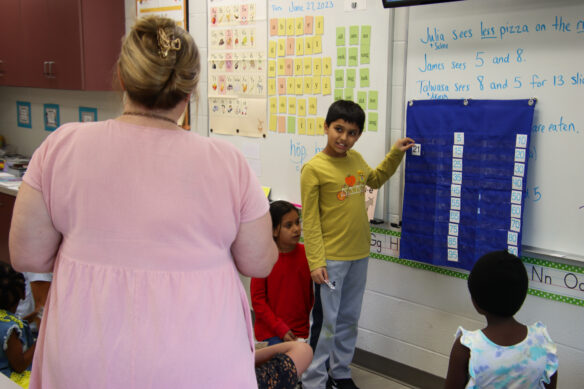 A child holds up a card that has the number 21 on it as he looks at a teacher in a classroom