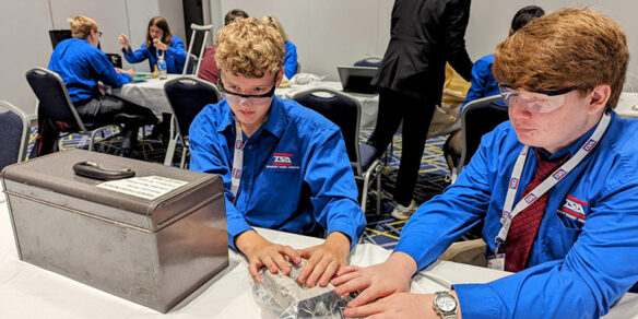 Two male students in matching shirts work on a piece of technology in a plastic bag on a table in front of them.