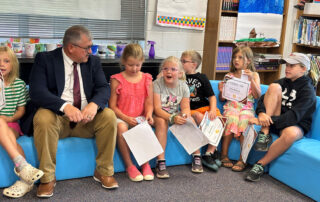 Larry Begley sits on chairs with young students in a classroom, talking to them.