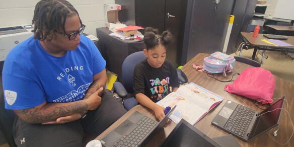 Seated at a desk in a classroom, an adult listens as a young girl reads from a book.