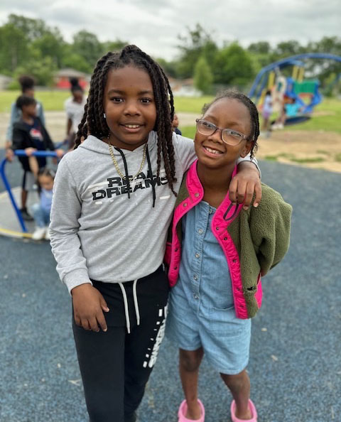 Two young students with their arms around each other smile for the camera while standing on a school playground.