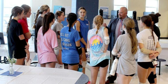 Willis Foster talking to a group of female volley ball players in a school cafeteria.