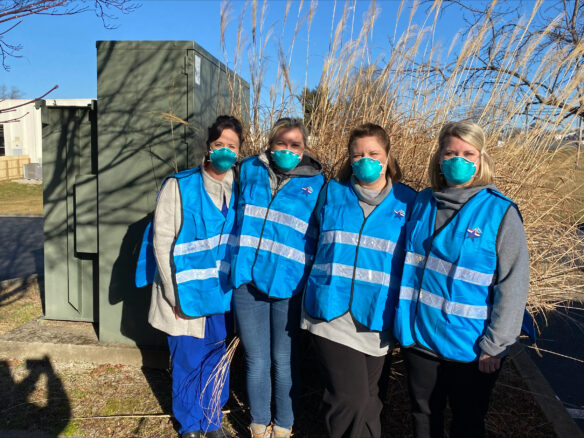 Four women nurses are standing next to each other outdoors. They are all wearing masks and vests.