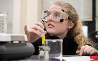 A female high school student wearing goggles kneels behind a desk with a test tube in her hand, surrounded by glassware.