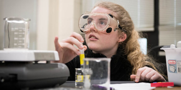 A female high school student wearing goggles kneels behind a desk with a test tube in her hand, surrounded by glassware.