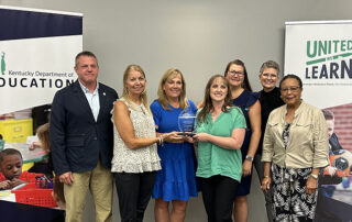 Jason Glass and Sharon Porter Robinson stand with a group of five smiling women who are holding onto a glass trophy.