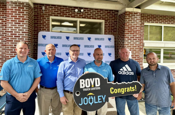 Six men stand in front of a school building holding a key-shaped sign reading: Boyd Truck Centers, Jouley, Congrats!