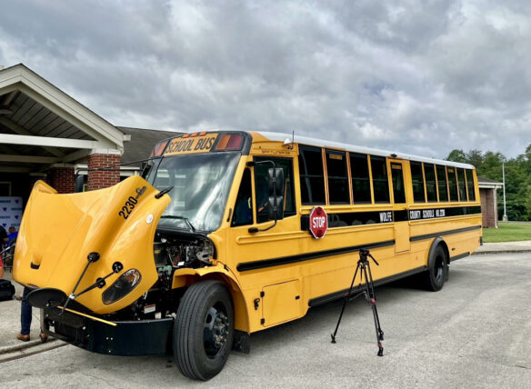 Picture of a bus with its hood up, sitting in front of a school building.