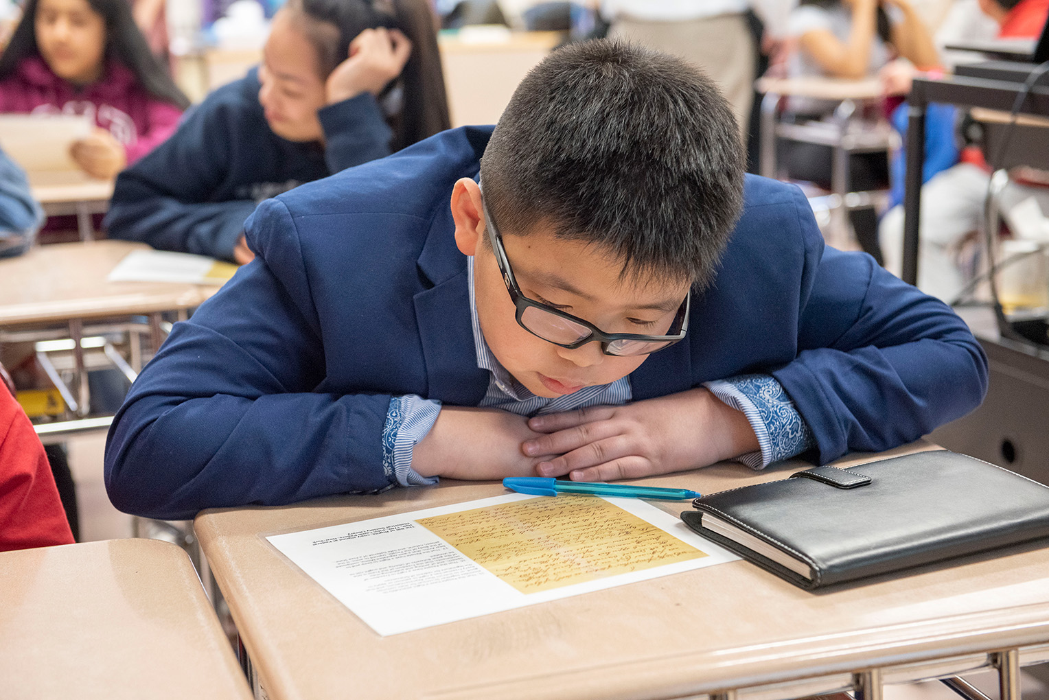 A kid reads text off a piece of paper while sitting at a desk