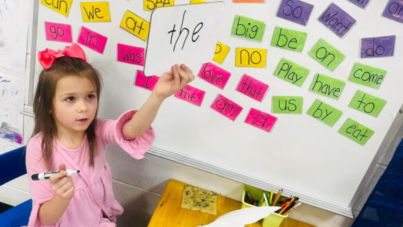 A child holds up a sign that says 'the' in front of a board with other words on it