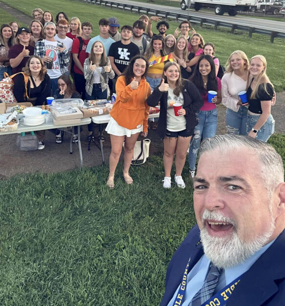 A group of smiling students at a buffet table stand behind Superintendent Todd Neace as he takes a selfie.