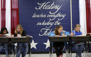Four students reading at their desks. Behind them is a red, white and blue wall with the saying, History teaches everything, including the future.